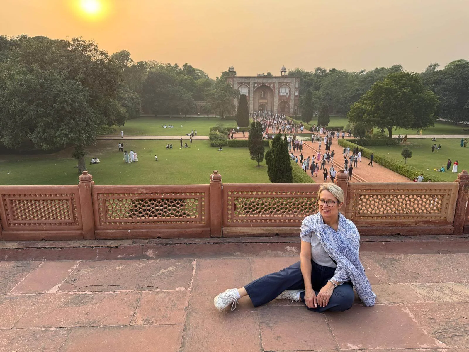 A woman sitting on stone pavement with a railing behind her, overlooking a lush green park with many visitors walking and relaxing, and a large historic building in the background during sunset.