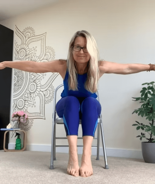 A woman with glasses and long blonde hair in a blue workout outfit seated on a chair with arms extended to the sides, performing a stretching exercise in a room with a patterned wall, a small table with flowers, and a potted plant.