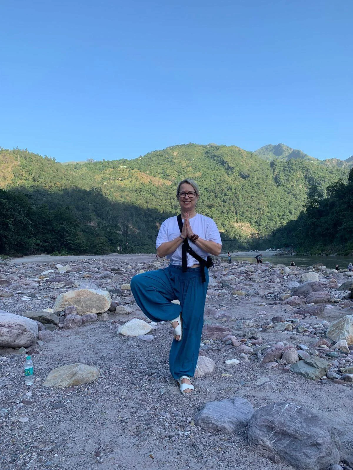 A woman practicing yoga by a river in a mountainous area, balancing on one leg with hands in prayer position, wearing a white top, blue pants, and white sandals.