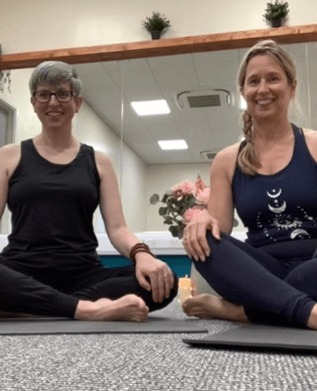 Two women sitting cross-legged on yoga mats in a peaceful indoor setting, smiling at the camera. One woman has short gray hair and glasses, wearing a black tank top. The other has long blonde hair in a braid, wearing a navy tank top with moon and star designs. There is a large mirror behind them, with potted plants and flower arrangements visible.