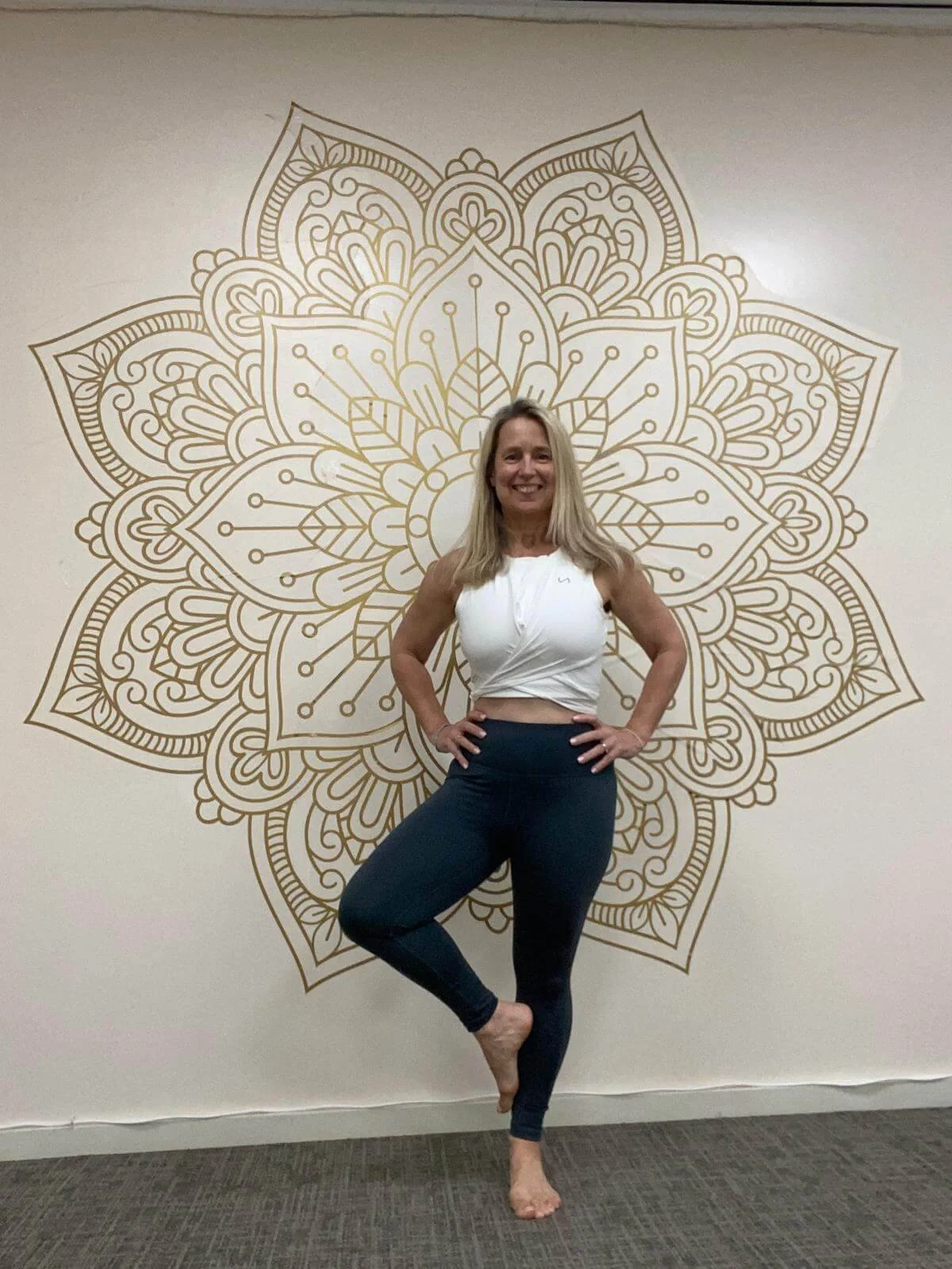 Woman practicing yoga indoors, standing on one leg with the other leg bent and foot resting on her thigh, in front of a decorative wall with a gold mandala design.