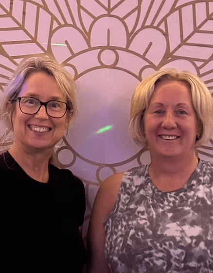 Two women smiling in front of a decorative pink and purple wall with a circular and fan-shaped pattern.