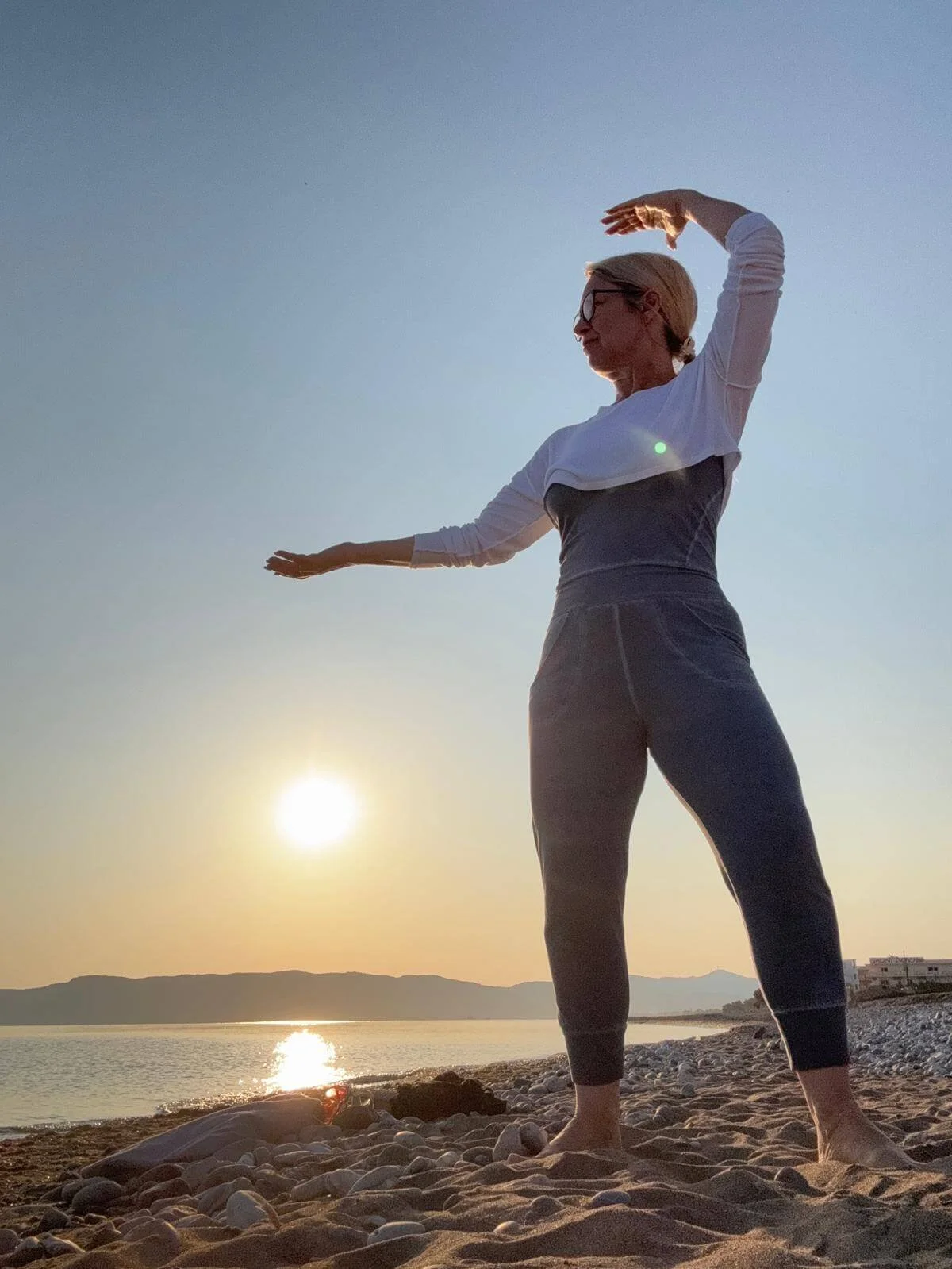 A woman practicing yoga on a beach during sunset, standing in a balanced pose with arms raised above her head and to her side.