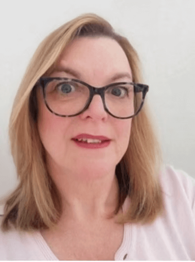 A woman with blonde hair, wearing glasses, and a light pink top, smiling at the camera against a plain white background.