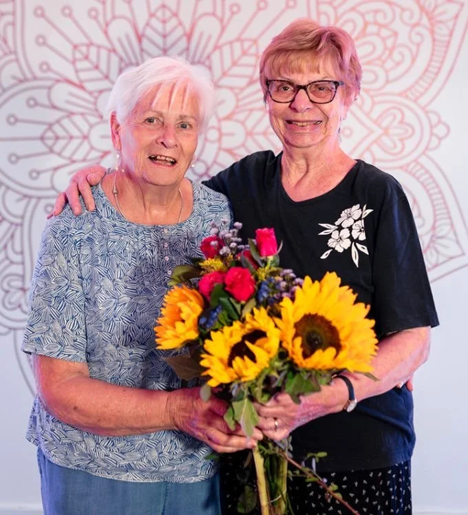 Two students of Discover Yoga Discover You smiling and holding a bouquet of sunflowers, pink roses, and other flowers.