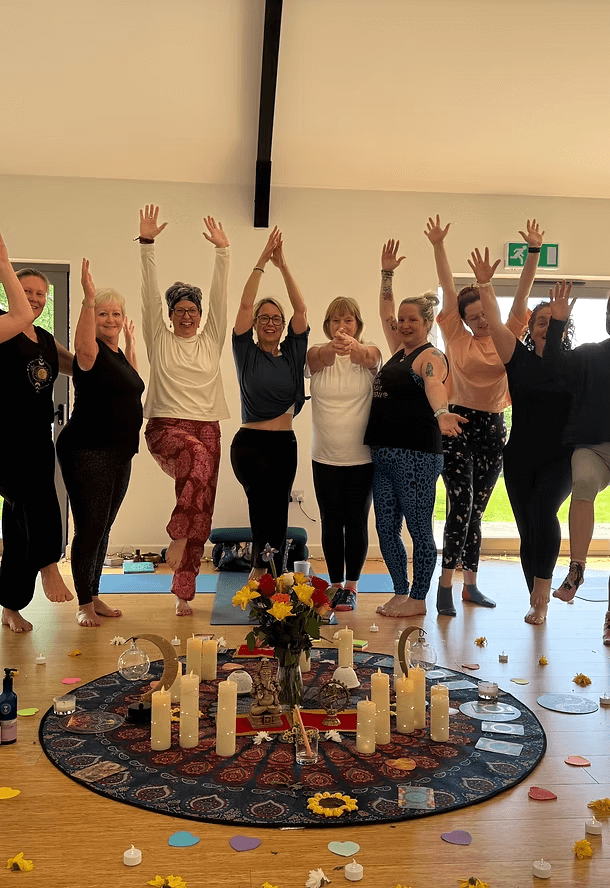 A group of women standing in a circle on a yoga mat, holding hands up and smiling inside a room with a circular shrine decorated with candles, flowers, and assorted objects; colorful paper hearts and flower petals are scattered on the floor.