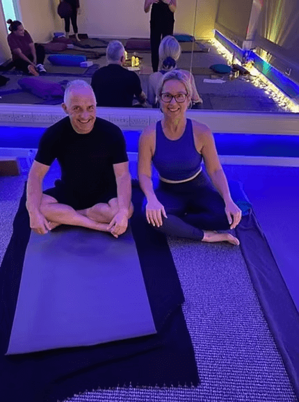 Two people sitting on yoga mats in a yoga studio, smiling, with others practicing yoga in the background and soft, colorful lighting.