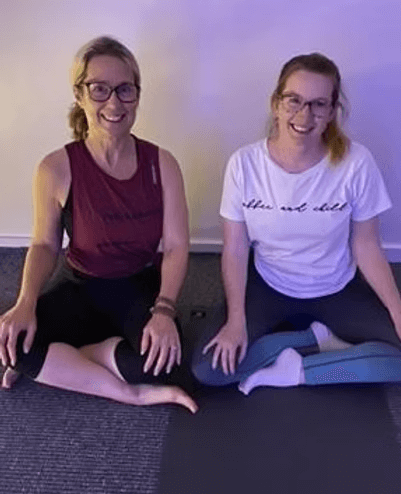 Two women sitting cross-legged on the floor, smiling at the camera, in a room with soft purple and yellow lighting.