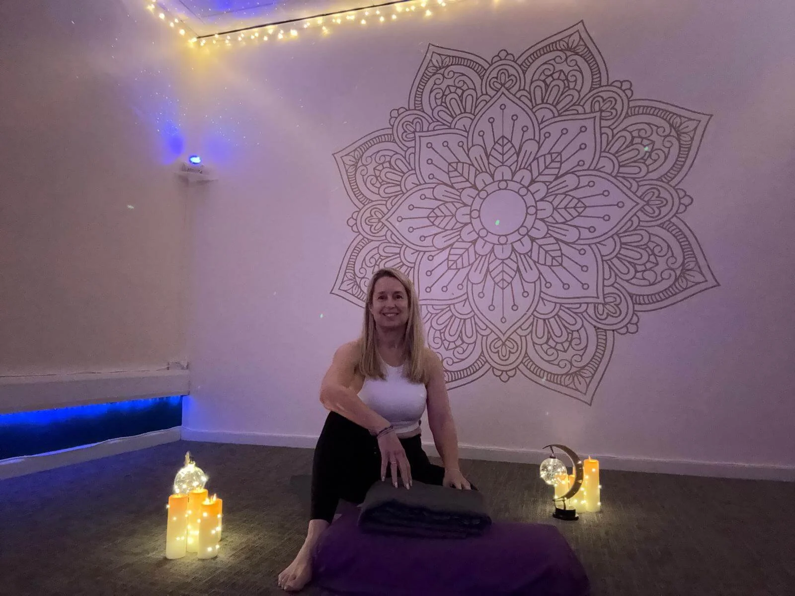 A woman practicing yoga in a serene studio with candles, fairy lights, and a large mandala wall art.