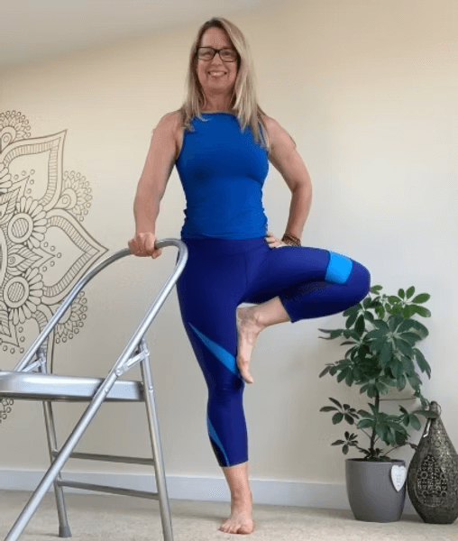 Woman in blue workout gear practicing yoga with one foot on a chair, balancing in a tree pose, smiling indoors.