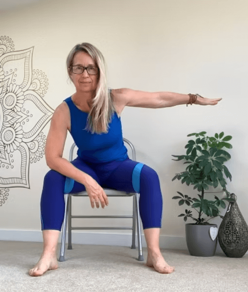 A woman wearing blue athletic wear sitting on a chair with one arm extended horizontally for a seated yoga pose or stretch, next to a potted plant in a modern room.