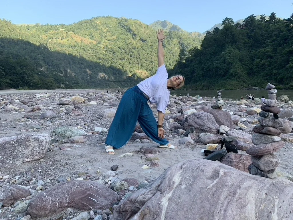 A woman practicing yoga on a rocky riverbank surrounded by mountains and forest, with stacked stones in the foreground.