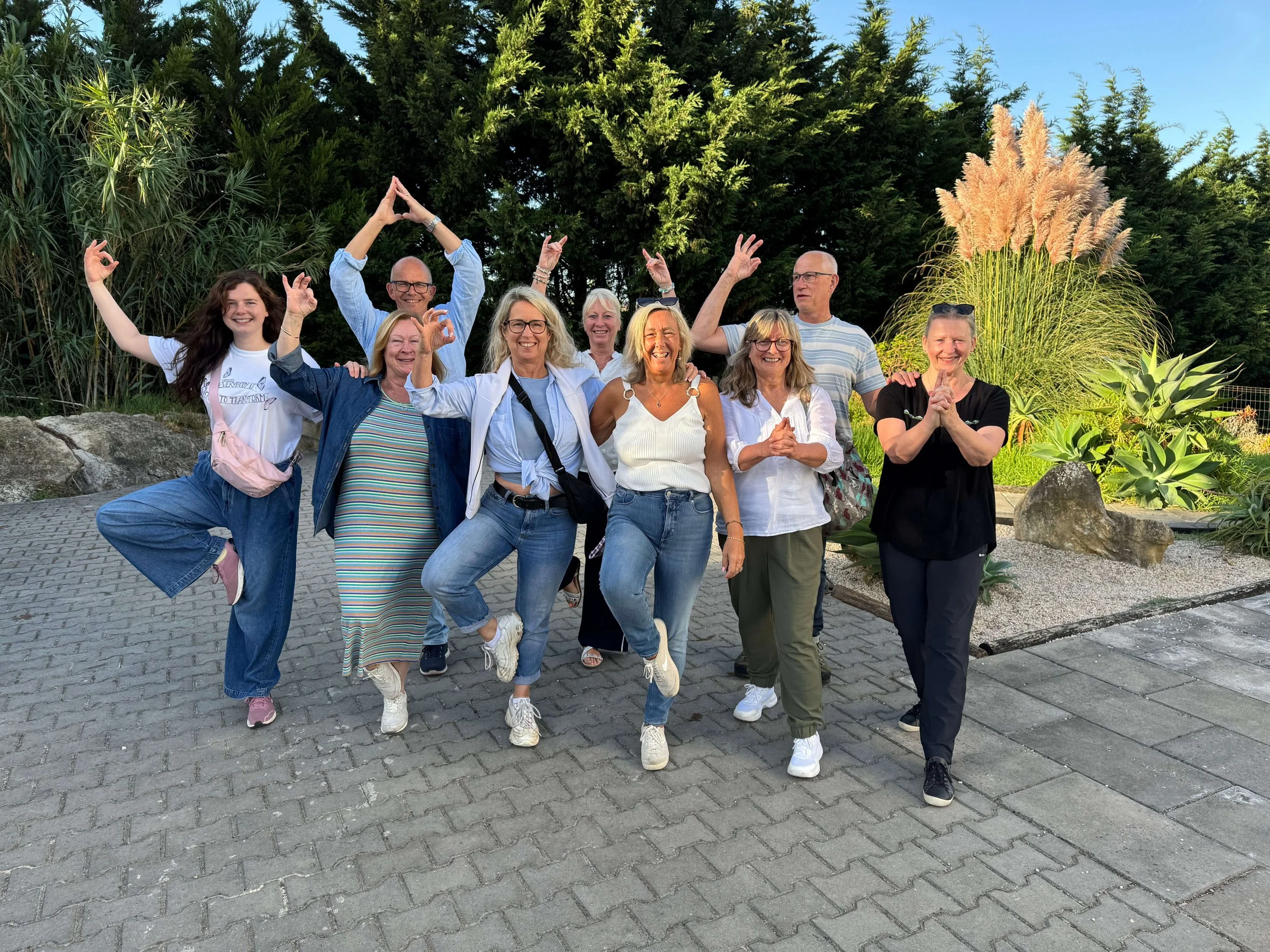A group of nine people, smiling and posing outdoors on a paved pathway with greenery in the background, some standing with one leg raised, some with hands raised or clasped.