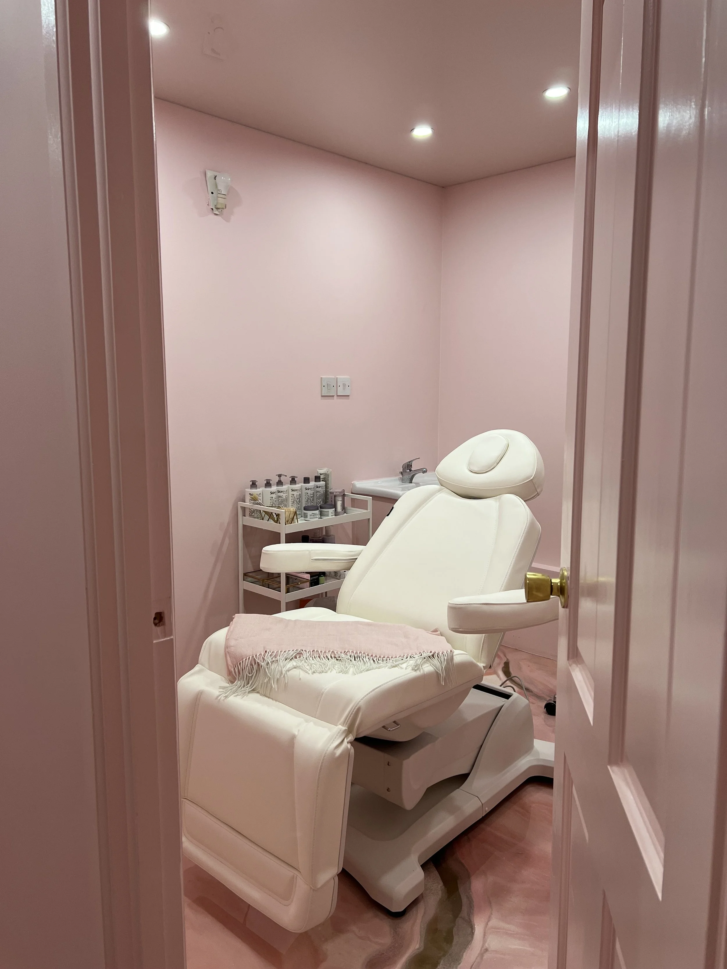 A pink-walled medical or spa treatment room with a white reclining chair, a small cart with bottles, and a sink.