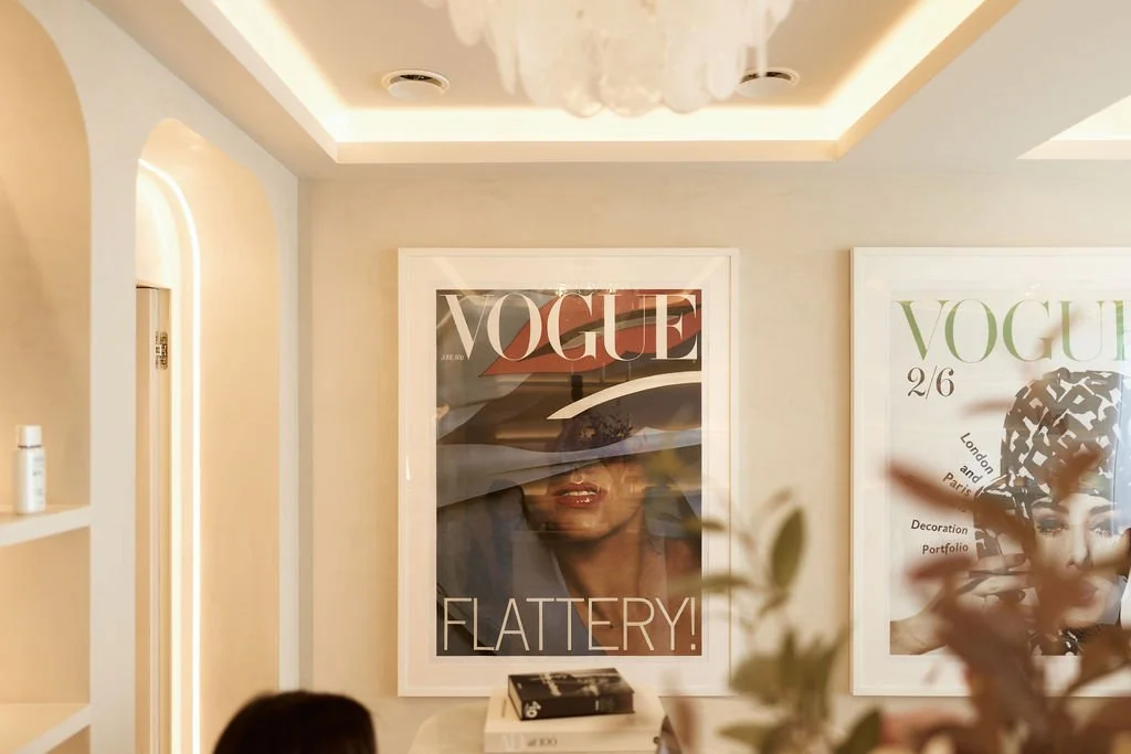 Interior room with beige walls and fashionable Vogue magazine covers hanging on the wall, one showing a woman with a wide-brimmed hat. There is a partial view of a person's head, a plant in front, and shelves on the left.