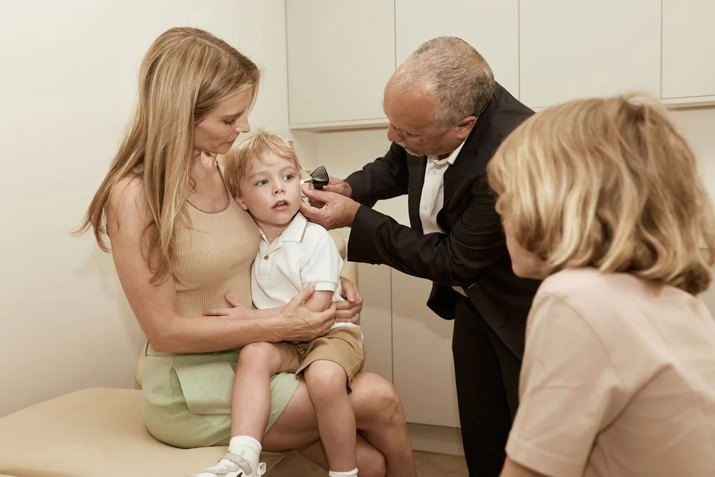A young boy sitting on a woman's lap at a doctor's office, seeing a doctor examining his ear with an otoscope, while a woman and a woman sitting nearby watch.