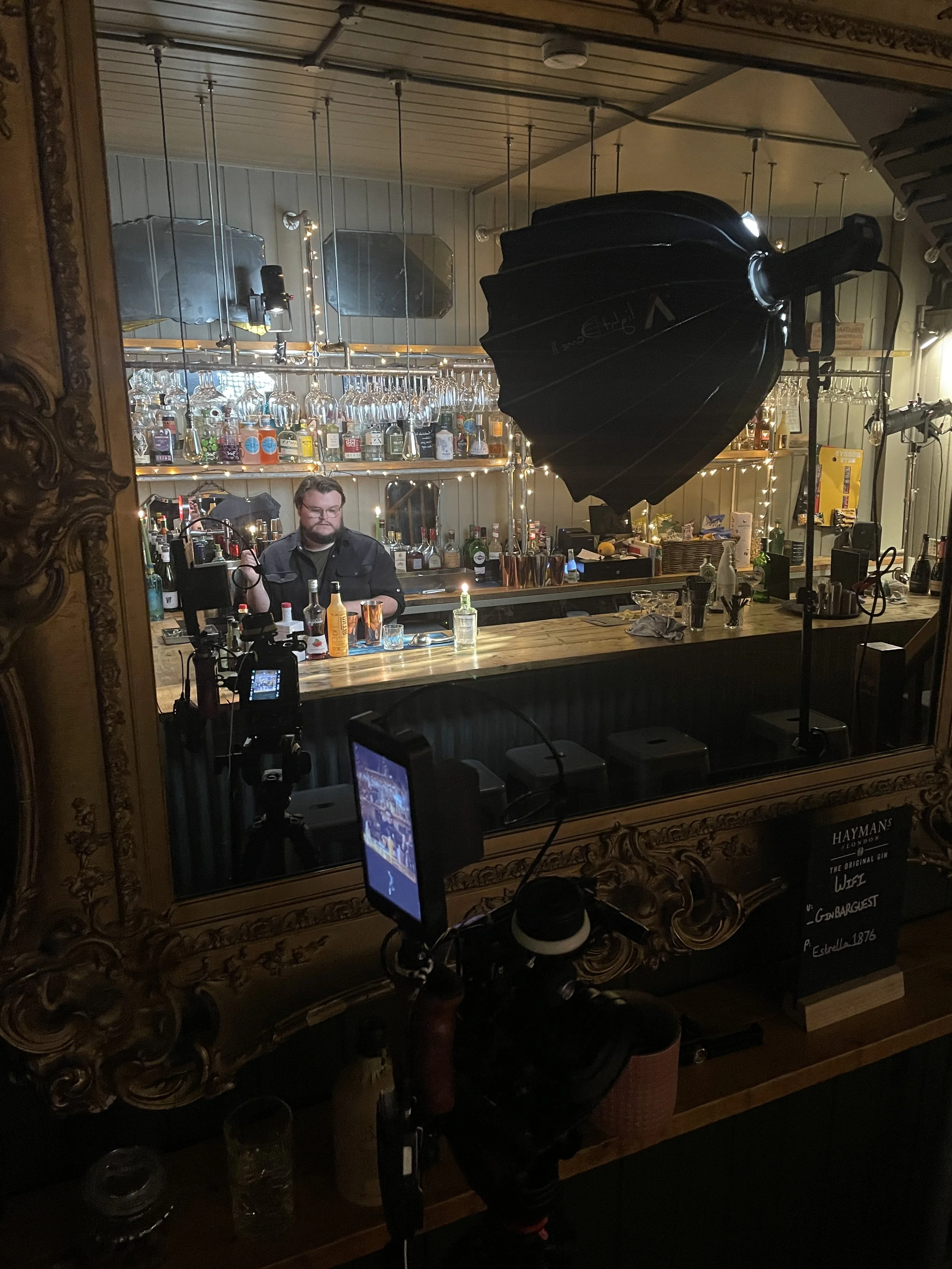 A bar scene with a bartender behind the counter, seen through a large ornate mirror. The bar is well-stocked with various bottles behind it and hanging glassware above. There is professional lighting equipment, including a large umbrella softbox, set up in front of the mirror, indicating a video or photo shoot.