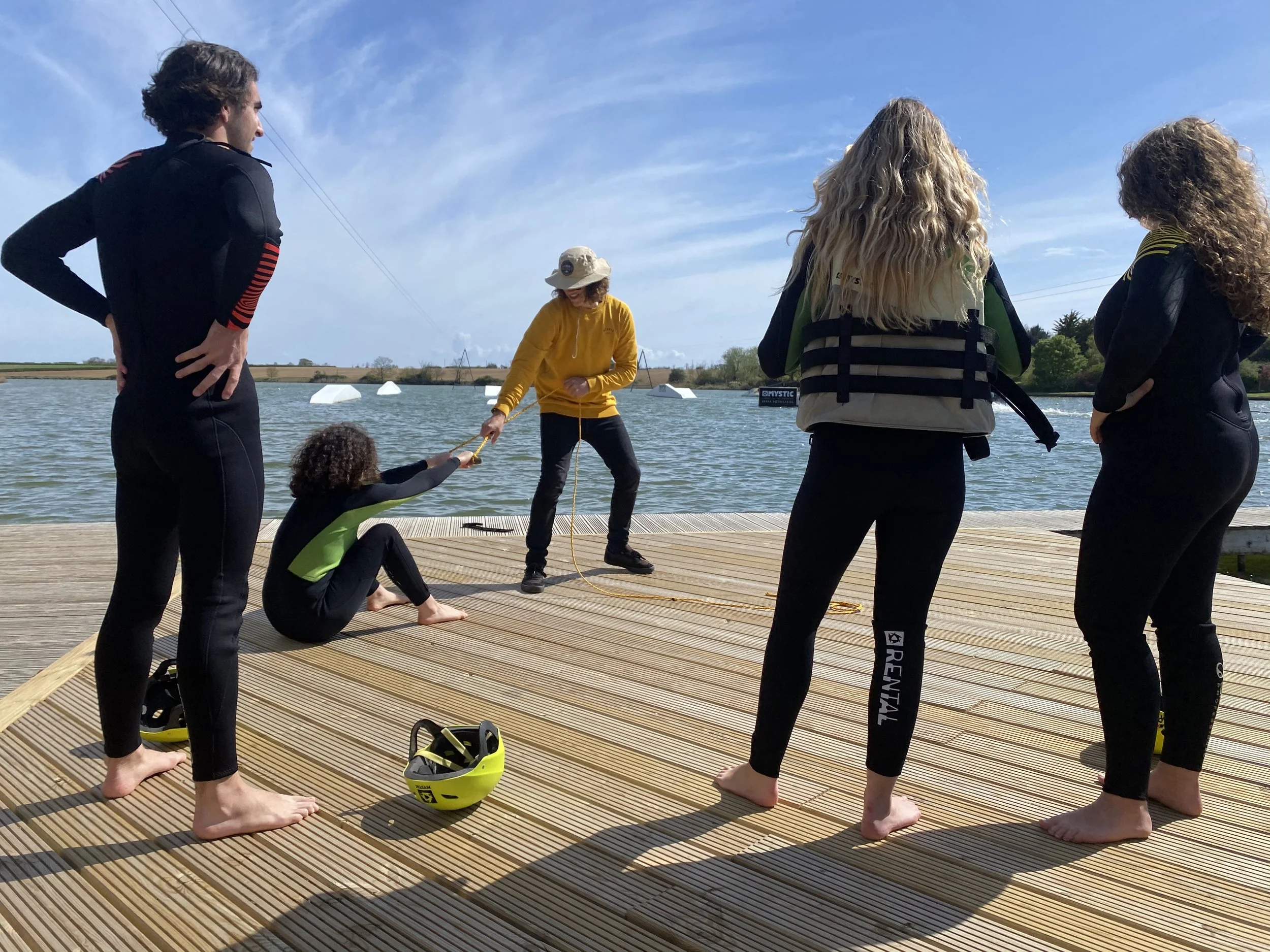 Group of five people, three women and two men, standing on a wooden dock by a body of water, participating in a beginners wakeboarding lesson at Curve Water Sports