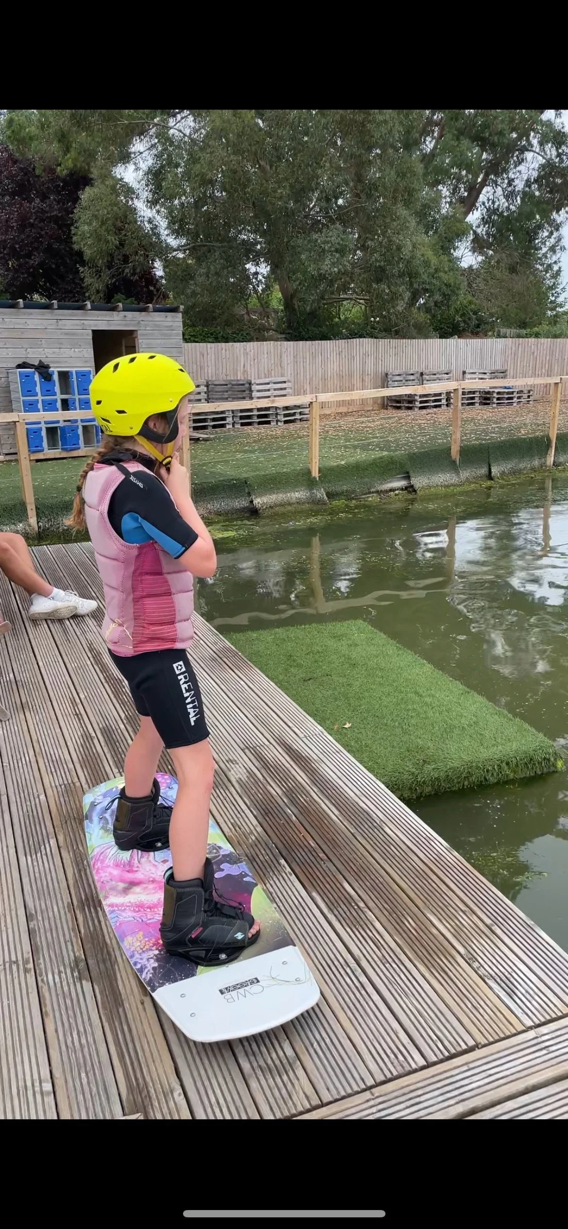 A young girl wearing a yellow helmet, pink life jacket, black wetsuit, standing on a wakeboard while participating in our kid's wakeboard club