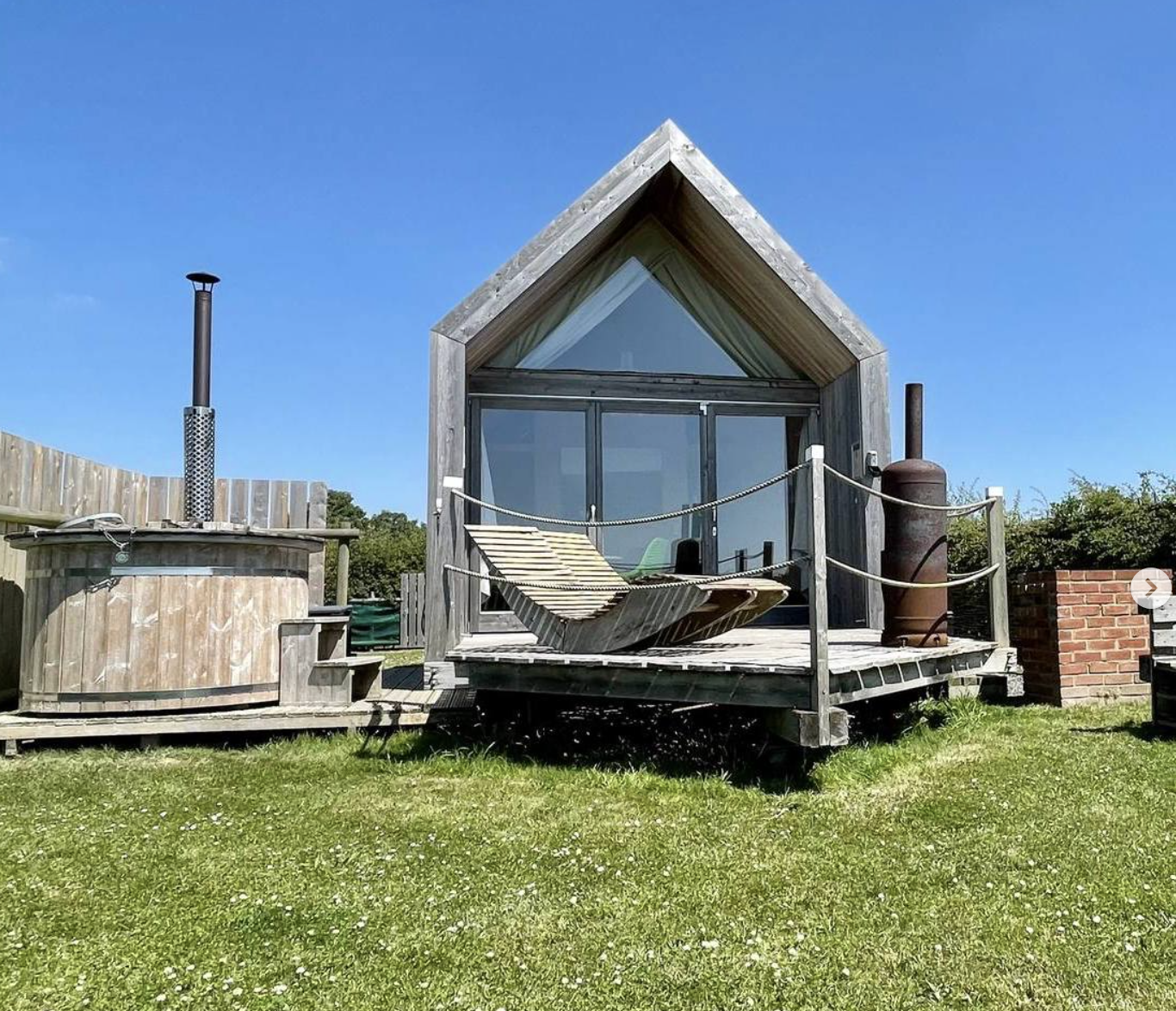 Lee Wick Farm Glamping Hut - A small wooden house with a triangular roof and large glass doors, situated on a grassy lawn, with a wooden lounge chair, a hot tub, a cylindrical metal heater, and a brick pillar nearby, set against a blue sky.