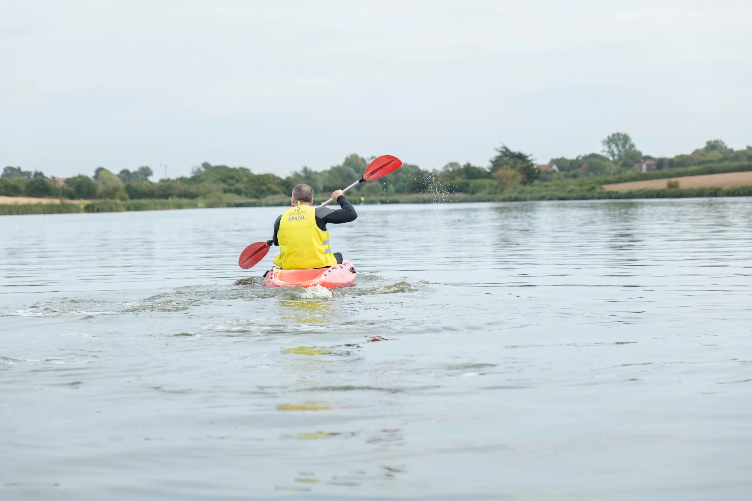 Person kayaking on a calm lake at Curve Water Sports Essex with green trees and fields in the background.