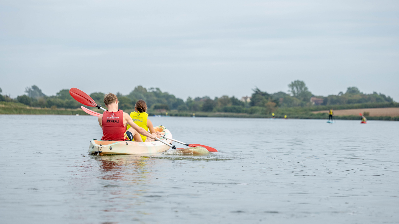 Two people kayaking on a calm river, with others in the distance, surrounded by trees and cloudy sky.