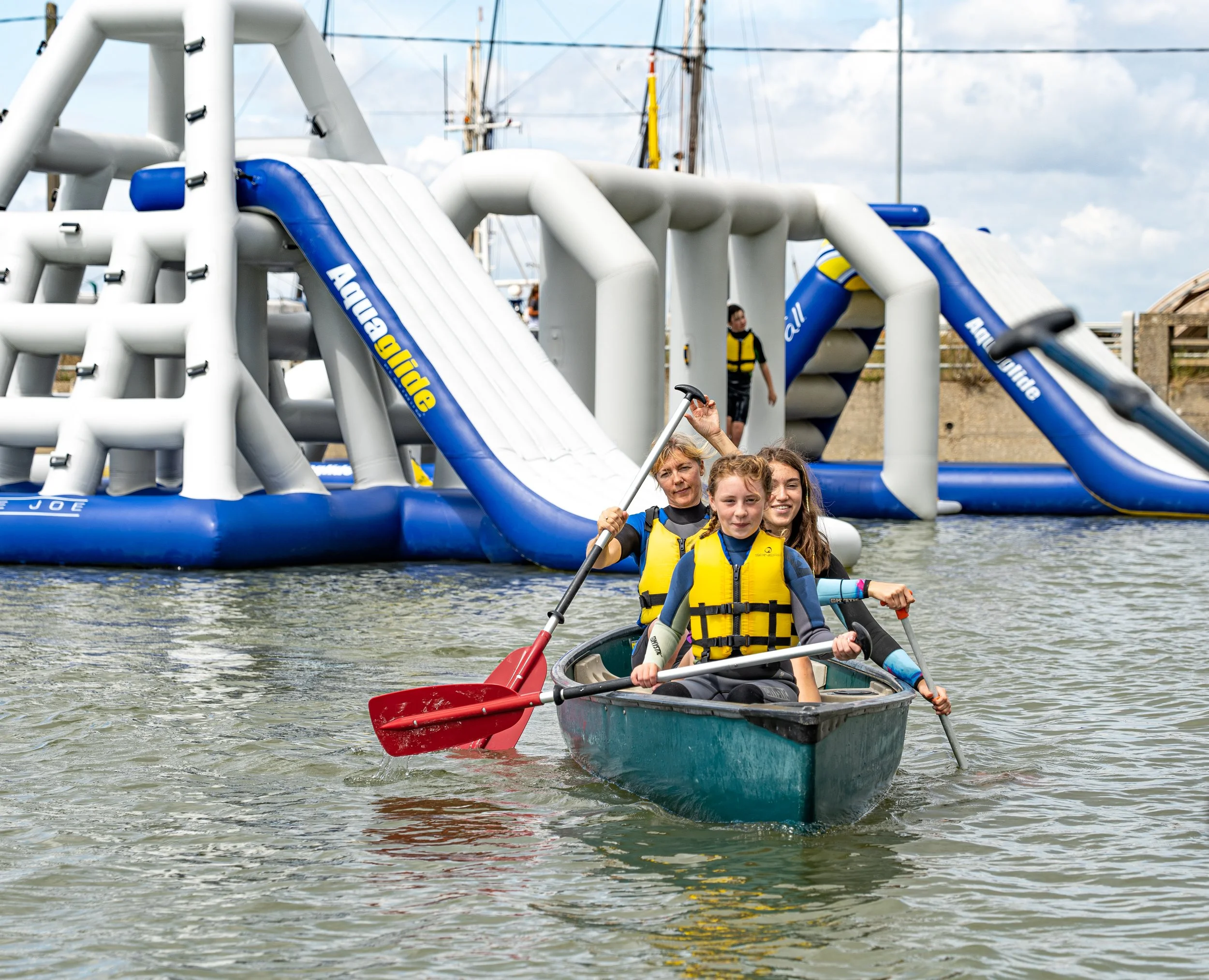 Three people in a kayak with life jackets, paddling on a body of water at Curve Water Sports Essex near an inflatable water park.