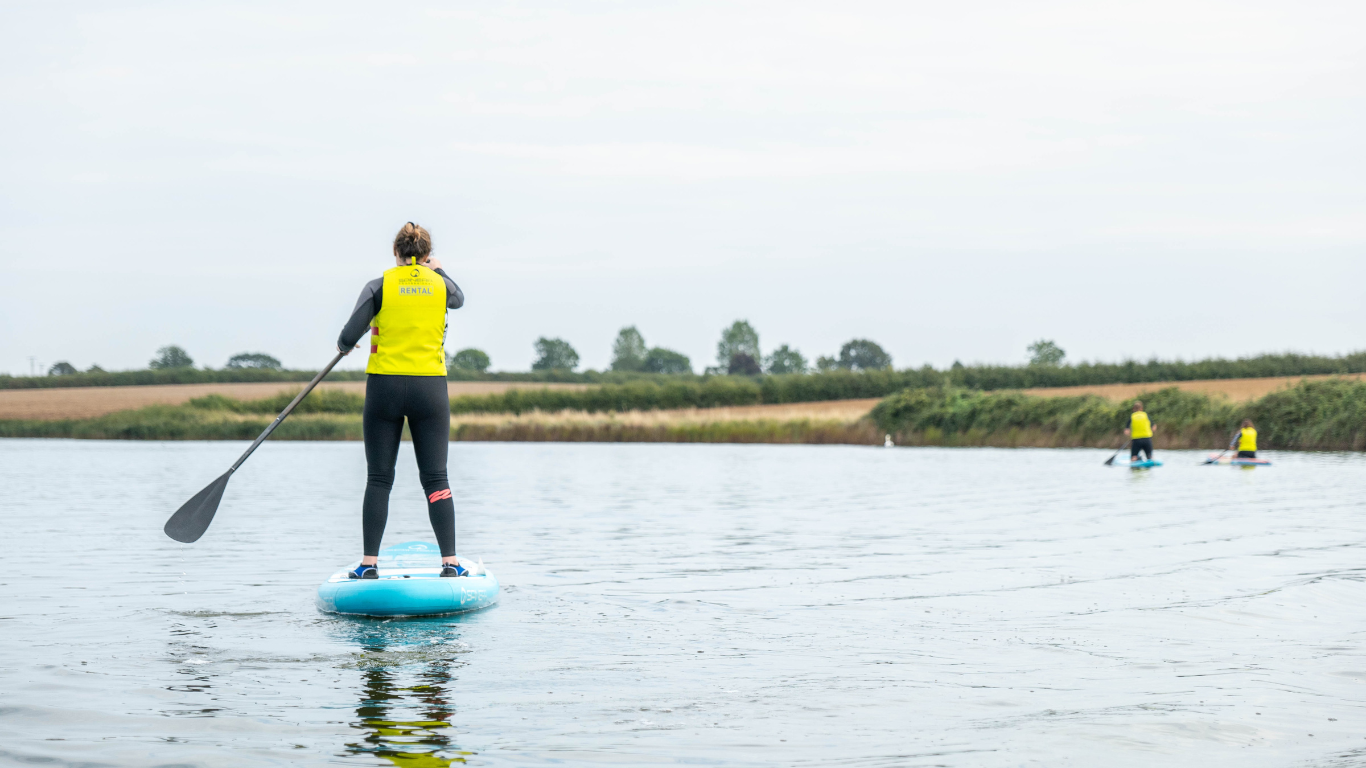 Person paddleboarding on a calm lake with two others paddleboarding in the background, all wearing yellow safety vests.