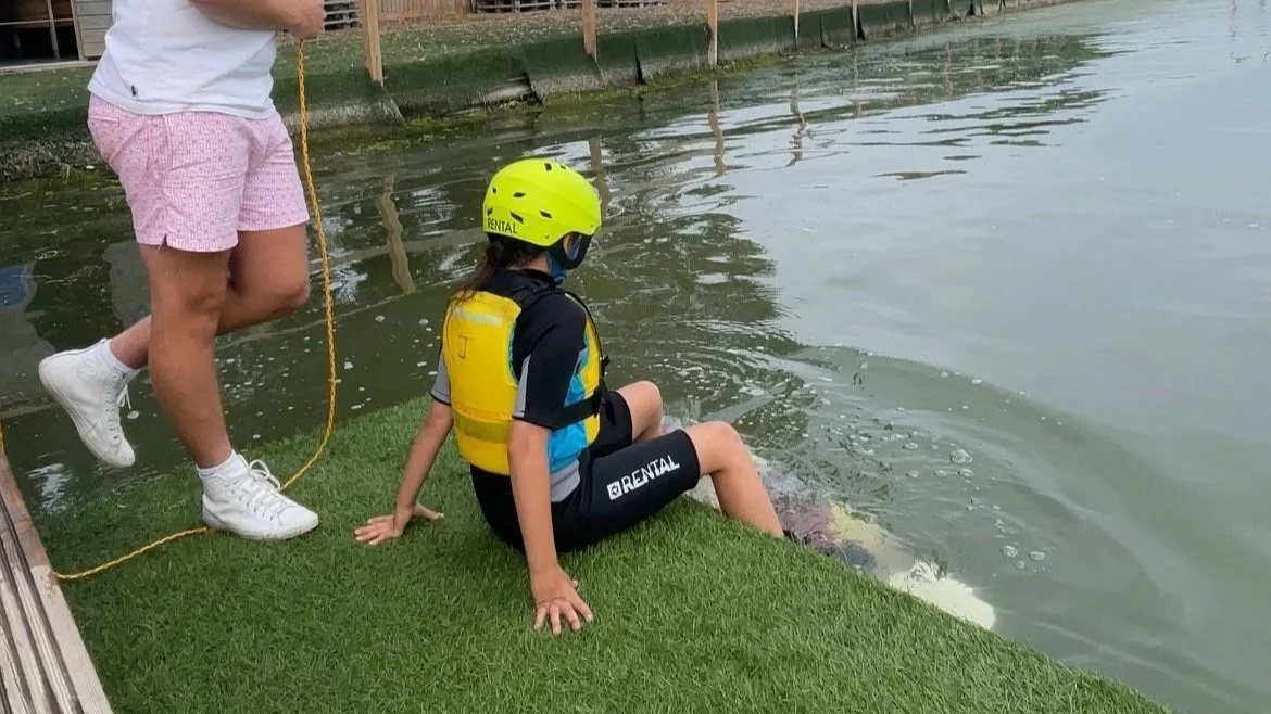 A child wearing a yellow helmet, life jacket, and black wetsuit sitting on a wakeboard dock by the water, while participating in our kid's wakeboard club.