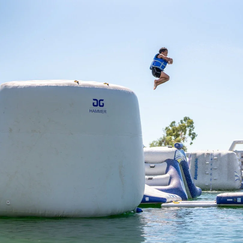 The 'Hammer' a large inflatable platform with a child mid-air jumping off the obstacle in a lake on a sunny day.