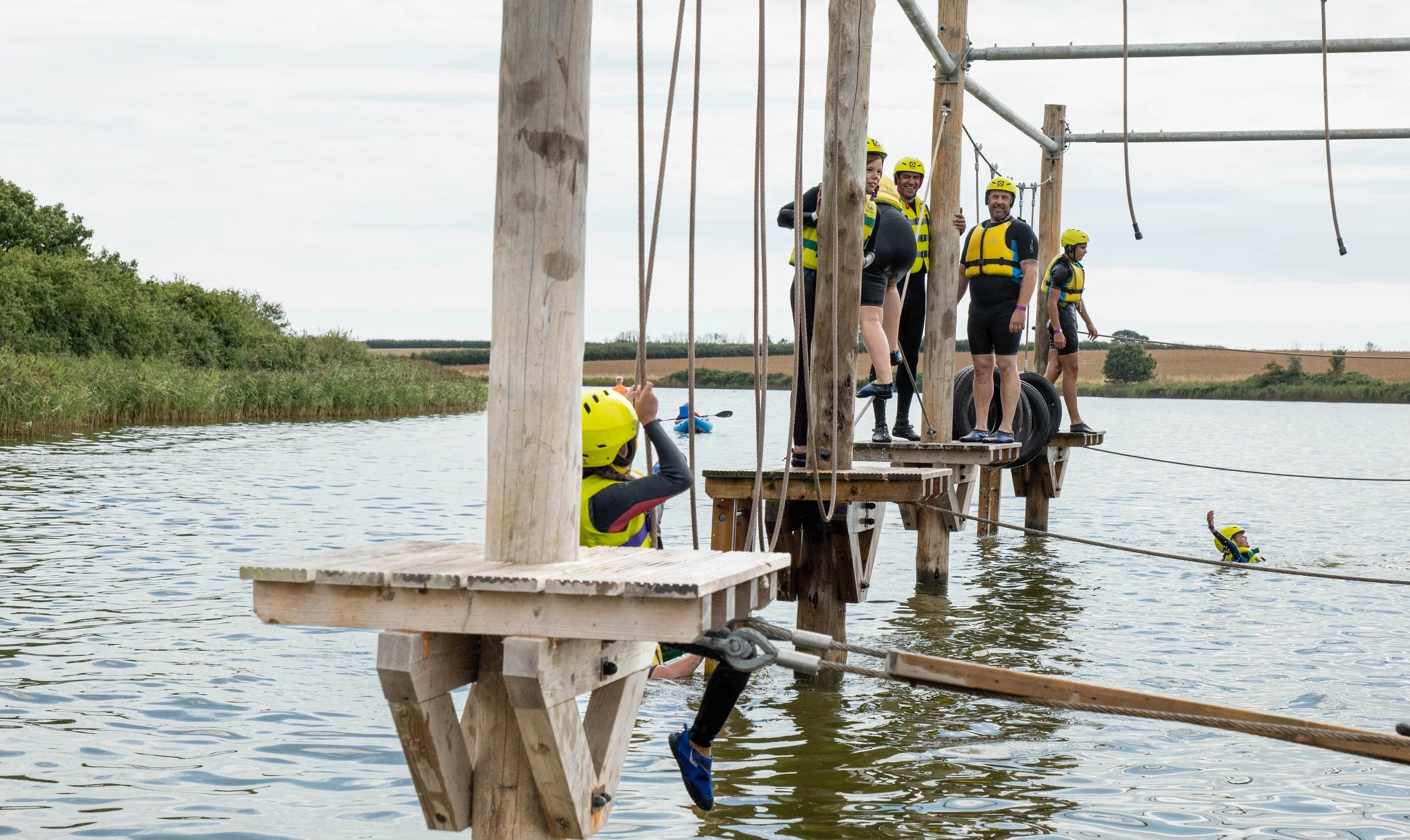 Group of people in yellow life jackets and helmets standing on our wooden Aqua Climb course over the water, preparing for zip lining adventure over a lake with trees and open fields in the background.