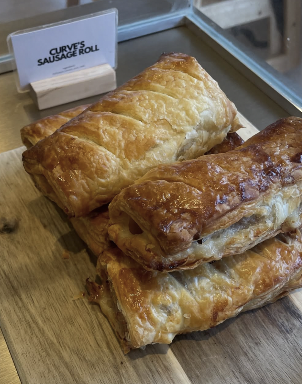 Three sausage rolls with golden, flaky pastry on a wooden cutting board, labeled 'Curve's Sausage Roll' in the background.