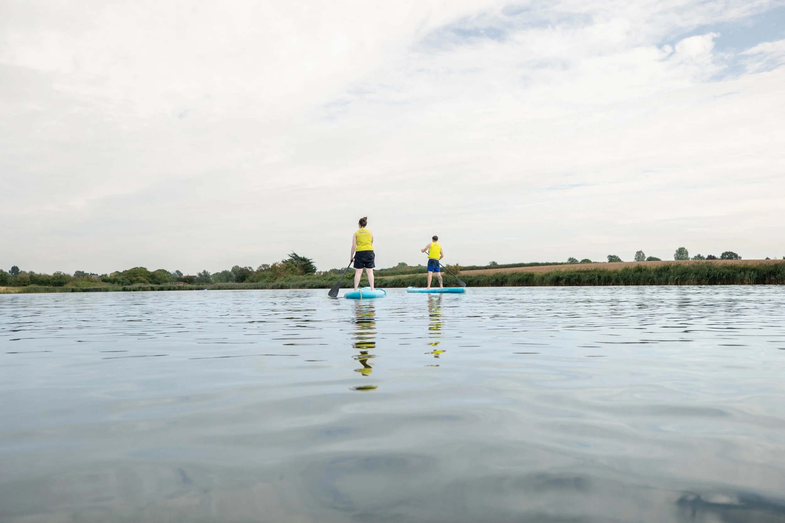 Two people paddleboarding on a calm lake at Curve Water Sports Essex under a cloudy sky, with a landscape of trees and open fields in the distance.