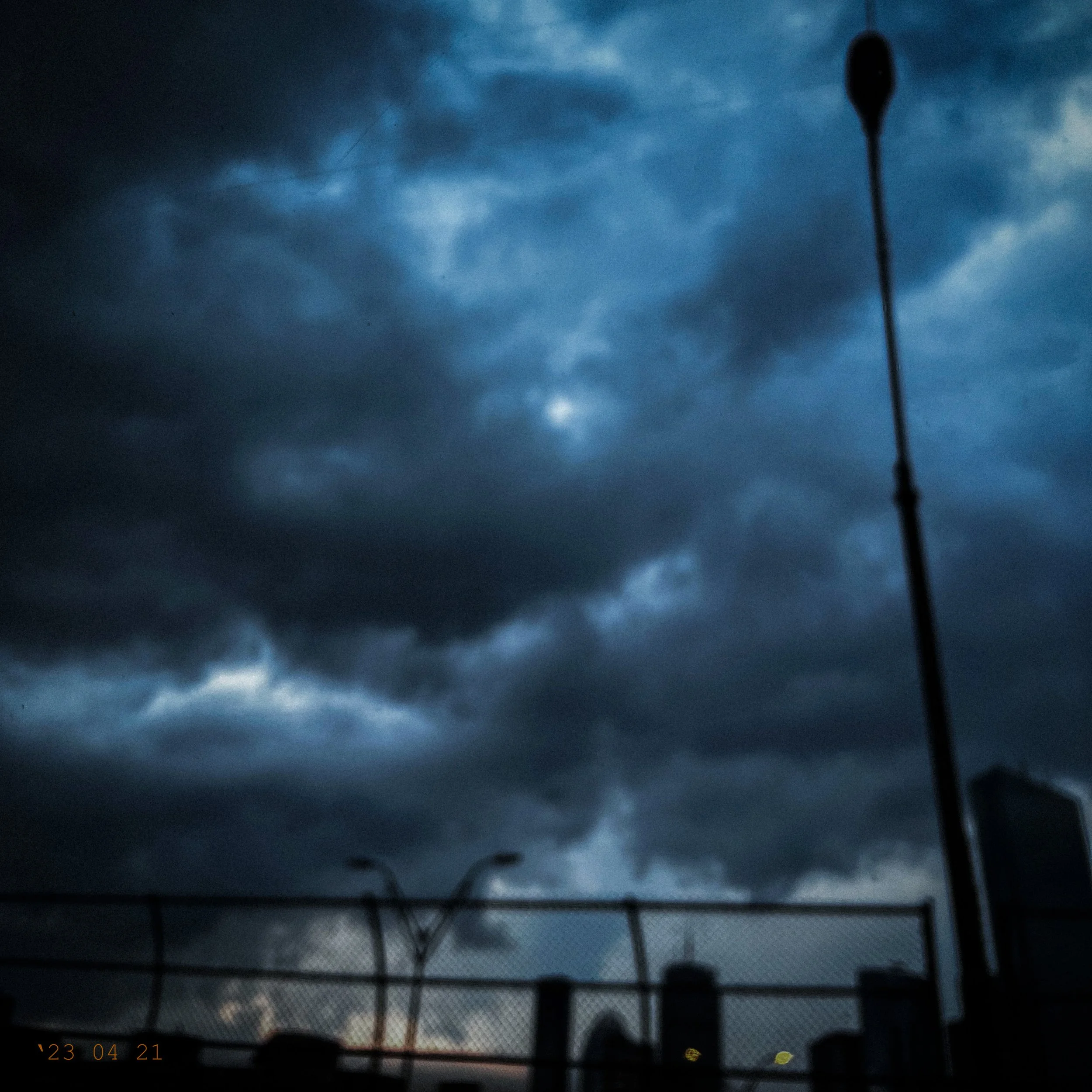 Dark cloudy sky over a cityscape with water towers and building silhouettes, with a chain-link fence in the foreground.