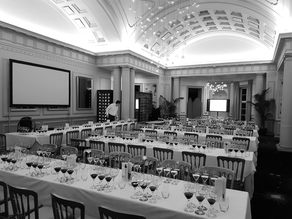 A banquet hall with multiple long tables covered in white tablecloths, set with wine glasses, water glasses, and tableware. The room has high ceilings with chandeliers, large windows, and decorative columns, and appears to be prepared for a formal event or tasting.