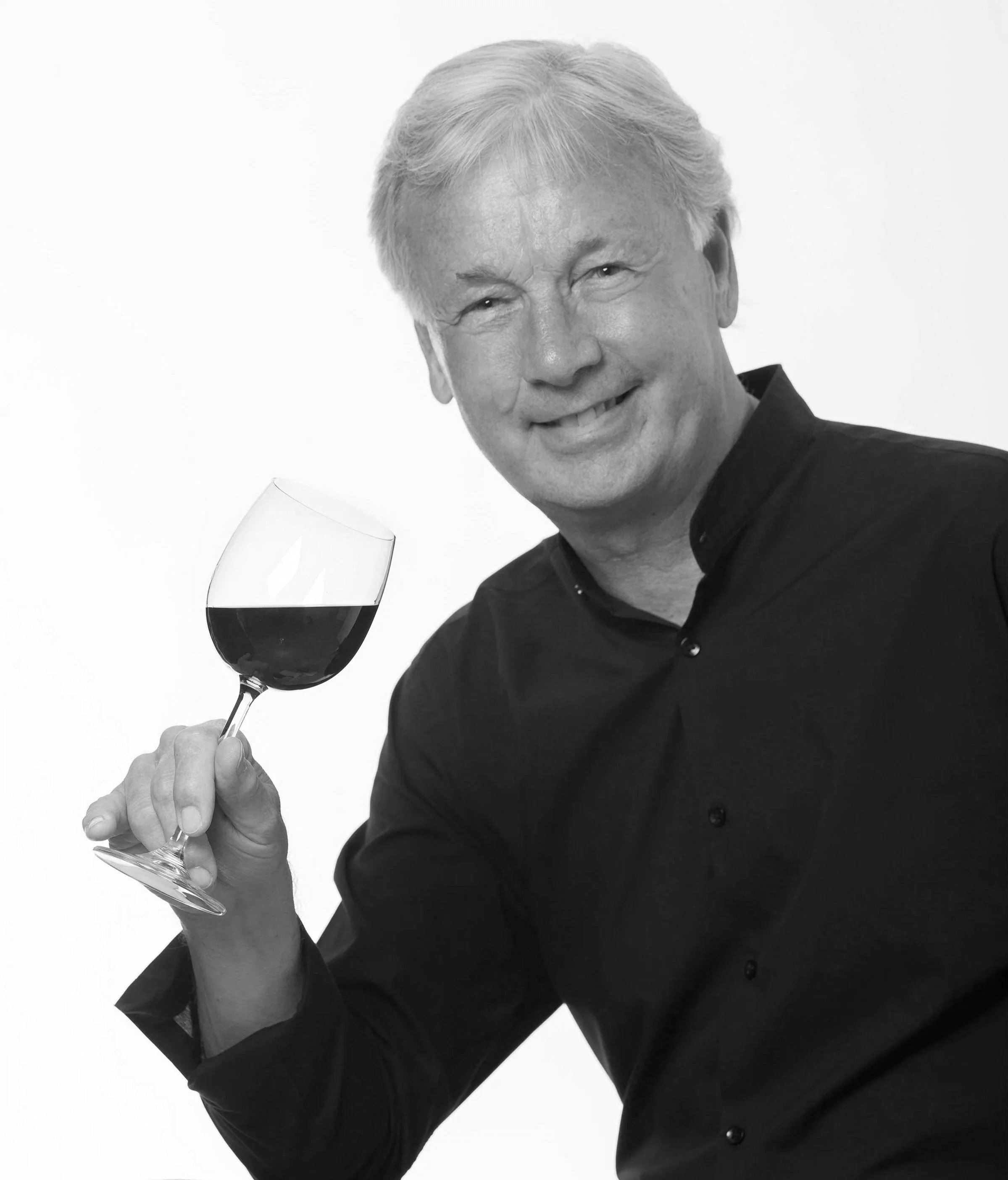 A smiling man with gray hair wearing a black shirt, holding a glass of red wine, portrait in black and white.
