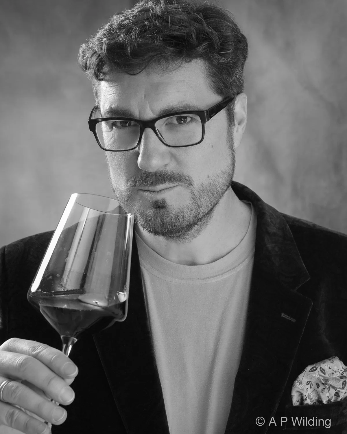 A man with glasses holds a glass of red wine close to his face, looking at the camera in a black-and-white photo.
