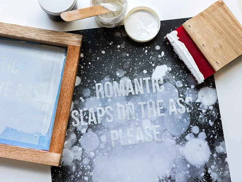 A stencil with the humorous message 'Romantic slaps on the ass please' on a black surface with white paint splatters, surrounded by wooden and art supplies.