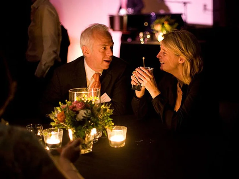 A man and woman sitting at a dimly lit table, talking and smiling. The man has gray hair and is wearing a suit and tie, while the woman has blonde hair and is holding a drink. There are lit candles and a flower arrangement on the table.