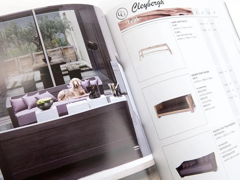 A magazine open to a page showcasing modern outdoor furniture, including a purple and white striped cushioned bench with a dog and glasses on it, next to a large window with a balcony and trees visible outside.