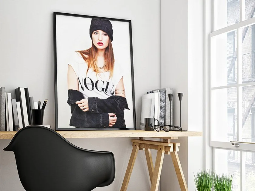 Interior of a modern home office with a large framed Vogue magazine cover featuring a woman in a black knit hat and black and white clothing, a wooden desk, black chair, and a large window letting in natural light.