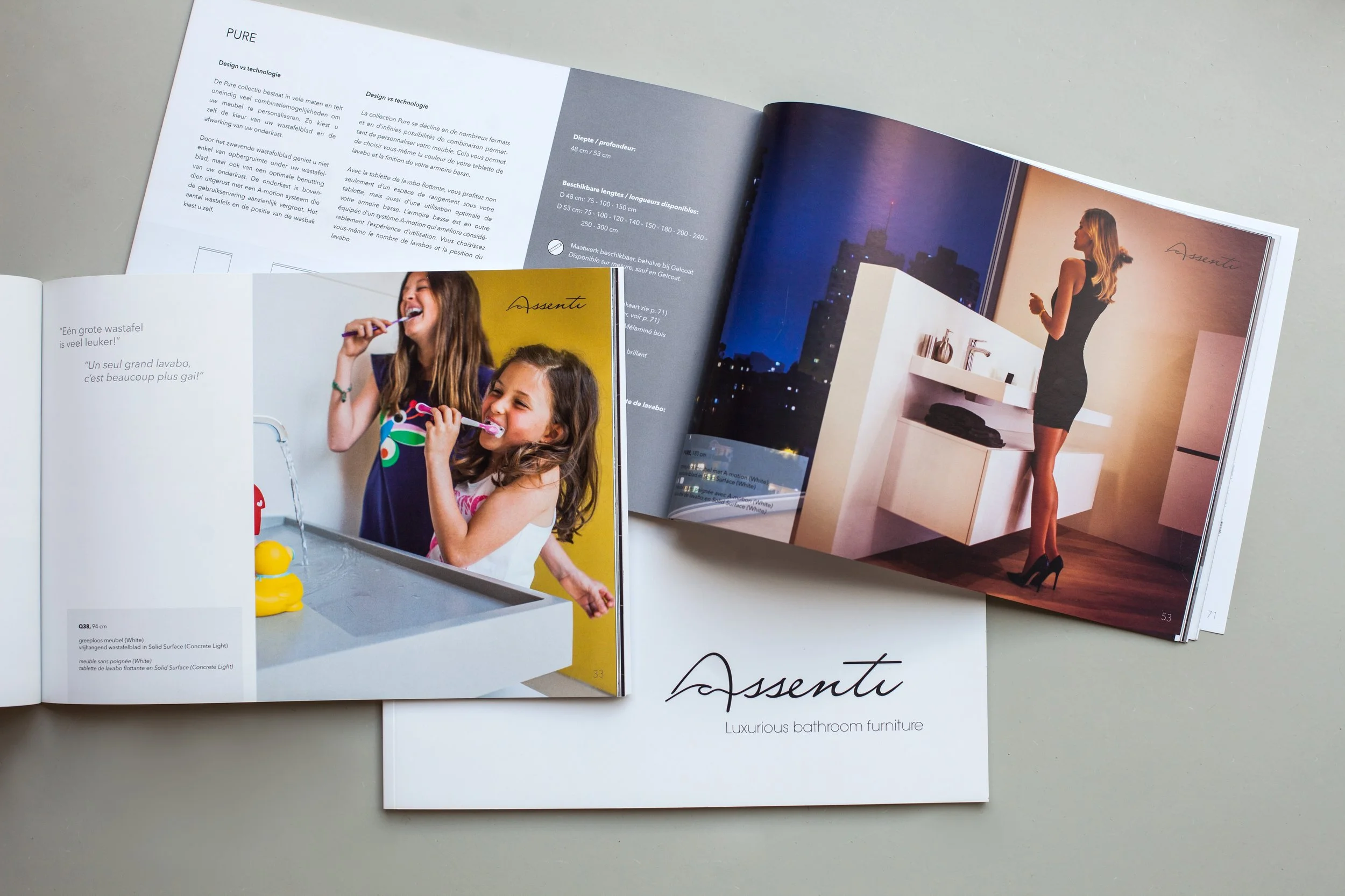 Open furniture catalog displaying images of bathroom furniture and lifestyle scenes, including two young girls brushing their teeth and a woman in a bathroom at night, with pages showing product information and branding for Assenti.