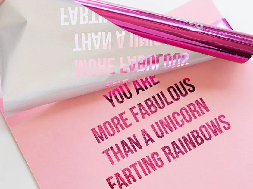 A pink sheet of paper with the message "You are more fabulous than a unicorn farting rainbows" written in bold pink letters. A mirror roller partially covers the text and reflects some of the message.