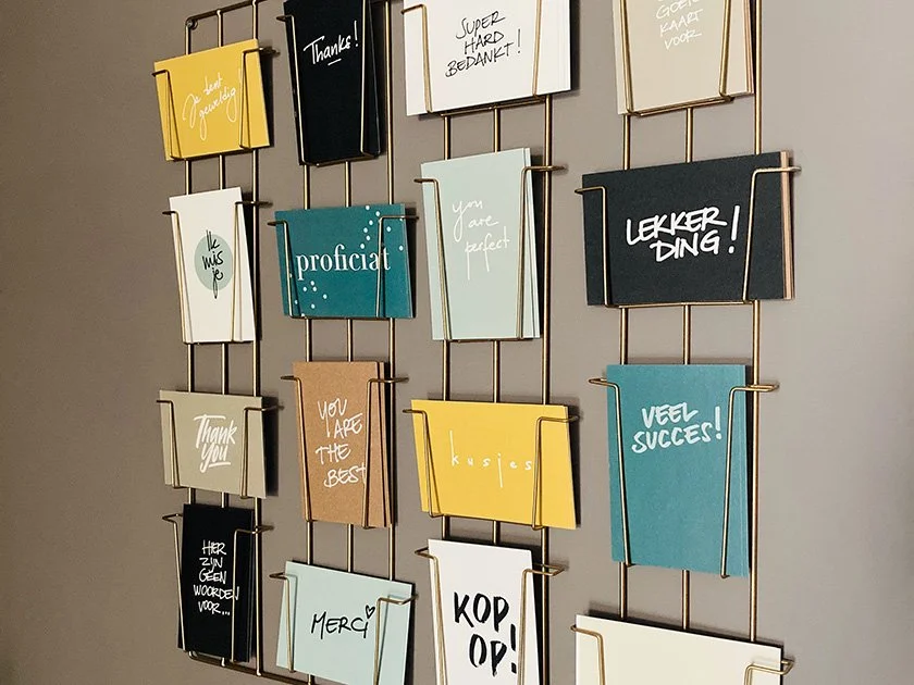 A wall display of greeting cards or notes with various messages, held in a metal grid. The cards are in different colors and styles, with handwritten and printed messages in Dutch and English, including 'Thanks!', 'You are perfect', 'Proficiat', 'Tha