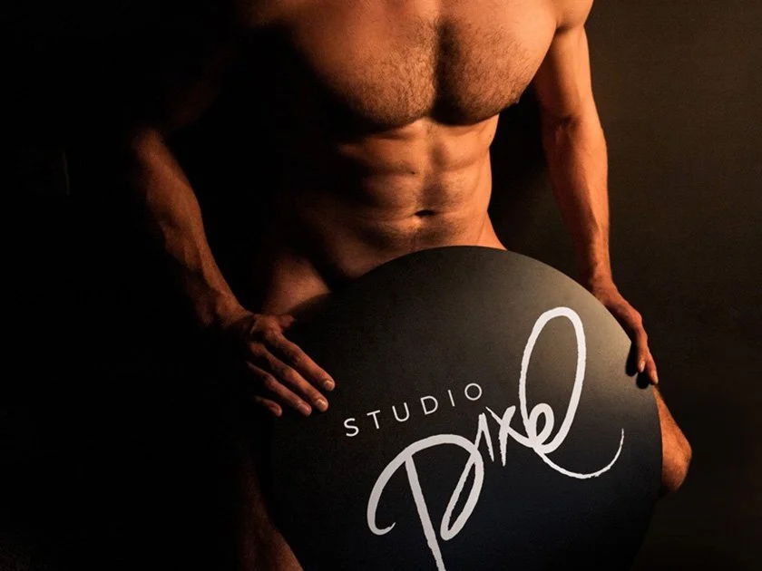 A muscular, shirtless man holding a black circular sign with the words 'Studio Dike' written on it, against a dark background.