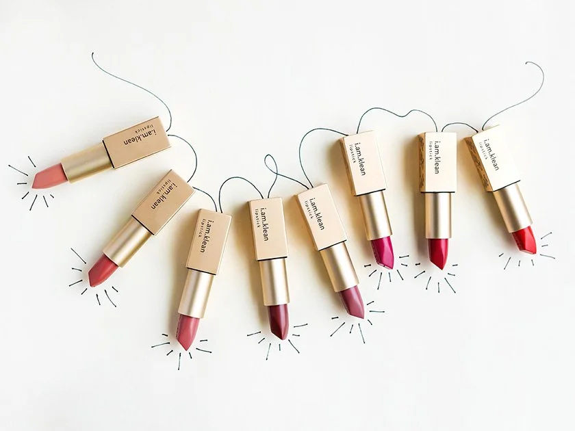 Seven lipsticks with beige caps labeled 'i am Kate' arranged on a white surface. Each lipstick has drawn lines around it resembling sparkling effects.