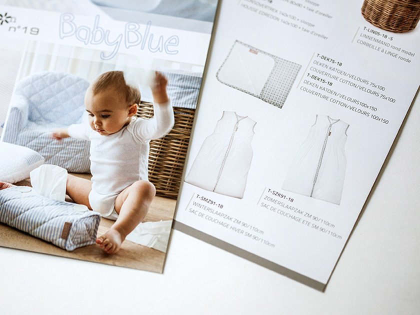 A baby sitting on the floor with a blanket, with a blue quilted pillow and a wicker basket nearby, and an open sewing pattern magazine showing dress design sketches and fabric details.
