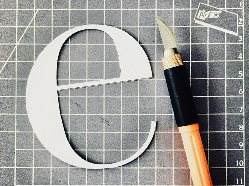 White paper letter 'E' on a gray cutting mat with a craft knife nearby.