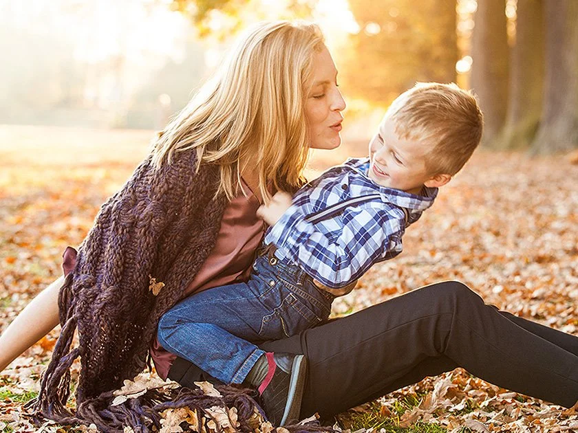 A woman and a young boy sitting outdoors on autumn leaves, playing and laughing together.