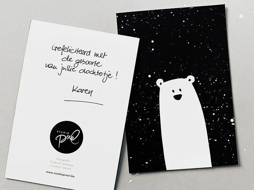 A greeting card with a handwritten message in Dutch and a black card with a white cartoon bear illustration surrounded by white splatters, resembling stars.